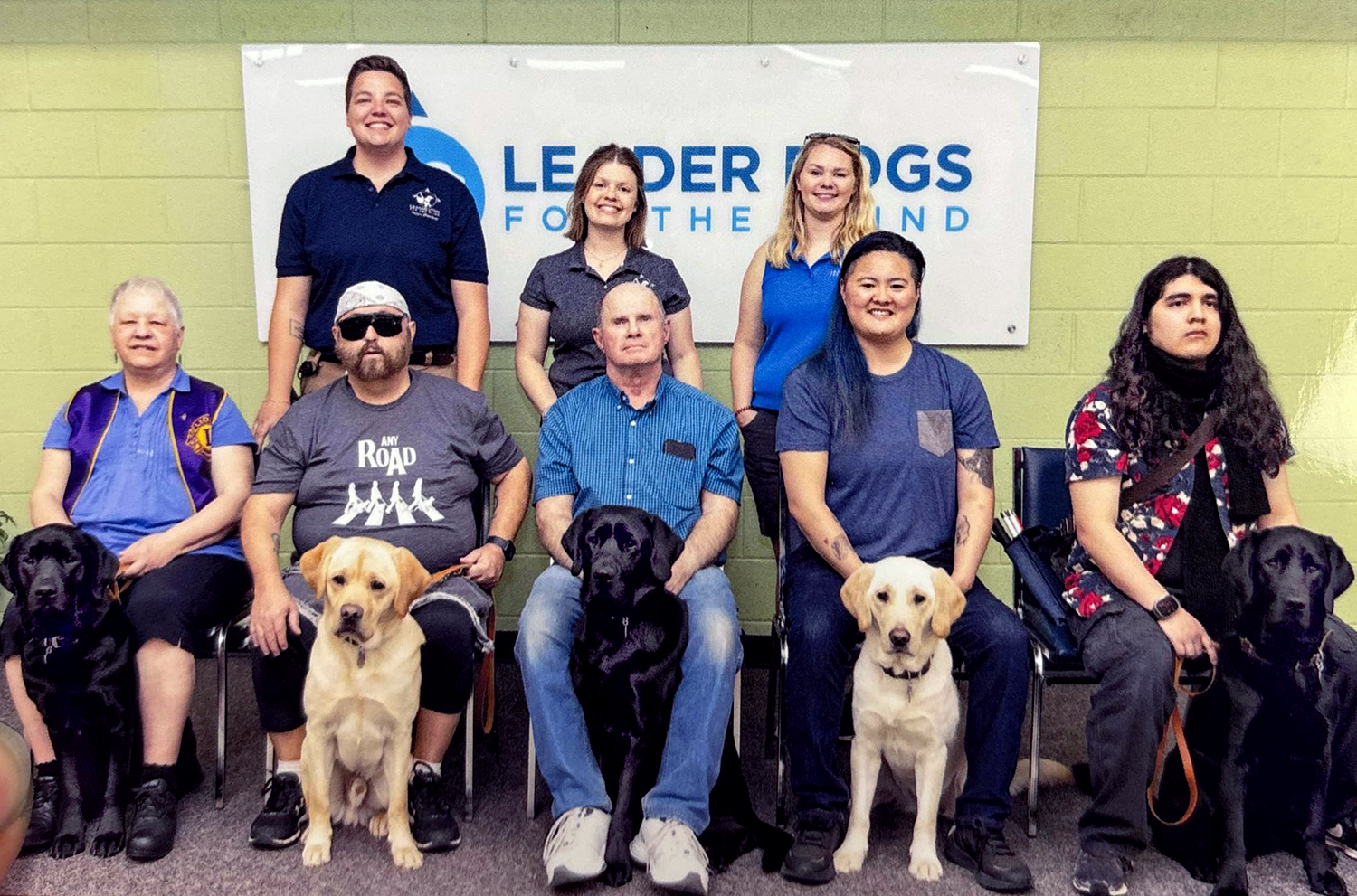 leader-dog-class This is a May 2025 class photo taken by Leader Dogs for Blind. The background wall has the Leader Dogs’ logo. The people in front of the wall are: Standing behind the clients siting in chairs are from your left to right: Chelsea, Emily and Mary. Sitting in row of chairs with dogs positioned between the clients legs looking from your left to right are: Ade Haugen from MN with “Orbit” a black Lab, Gordan from OK with “Bruce” a yellow Lab, Dave from UT with “Oby” a black Lab and sibling of “Bruce”, Chelsea from CA with “Jasmine” a yellow Golden/Lab Cross and E’mon from CA with “Shadow” a black Golden/Lab Cross and sibling of “Jasmine”.