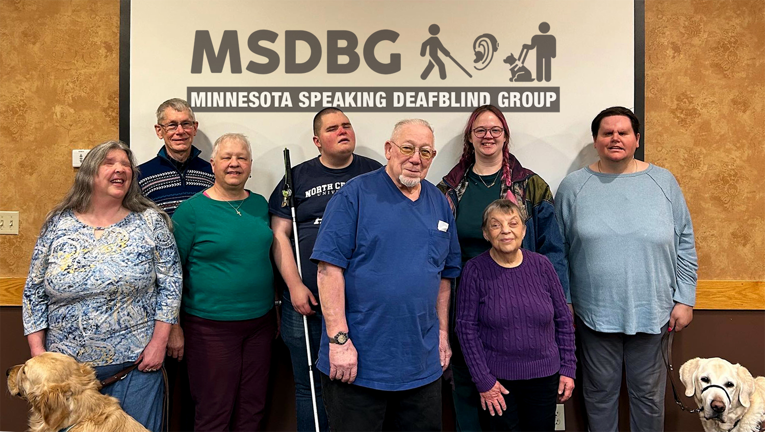 On the wall behind the retreat participants is the logo with uppercased letters of M S D B G and to your right of the letters are three graphic symbols going from left to right: a blind person walking with a white cane, a human ear with a hearing aid attached and a person standing while holding the leash of a guide dog sitting down. The people who were at the March 2025 MSDB Retreat are from left to right: in the back row are SSP Richard, SDB Rocky holding a white cane, SSP Grace, and SDB Hannah with guide dog “Ledger”. In the front row are SDB Tammy with guide dog “Makree”, SDB Ade, SSP/DLV Tom and SDB Suzanne.